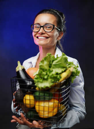 Portrait of a happy middle-aged woman in glasses and apron holds a basket of fresh vegetables and fruit. Studio photo on dark backgroundの写真素材