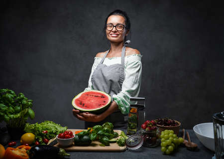 Middle-aged female chef holds half the watermelon in kitchen, smiling and looking on the camera. Healthy and proper nutrition on a diet. Studio photo on a dark backgroundの写真素材