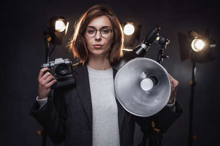 Portrait of a beautiful redhead female photographer holds a digital camera and looking on the camera. Studio shot on dark background with lighting equipmentの写真素材