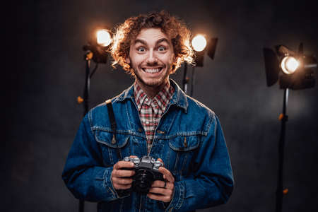 Funny handsome man with curly hair wearing denim jacket holds digital camera. Dark photo studio with lighting equipment in the backgroundの写真素材