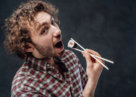 Emotional handsome man with curly hair holds chopsticks with sushi roll and looking on camera. Studio shot on dark backgroundの写真素材