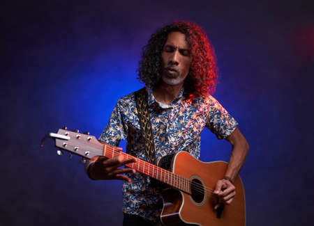 Middle aged hispanic man musician in a hawaiian shirt playing guitar on a dark illuminated by blue...