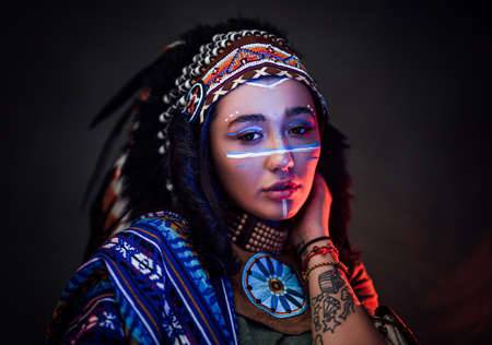 Charming American Indian girl with tattoo on hand in traditional make up hat and clothes on a dark background illuminated by red and blue lightの写真素材