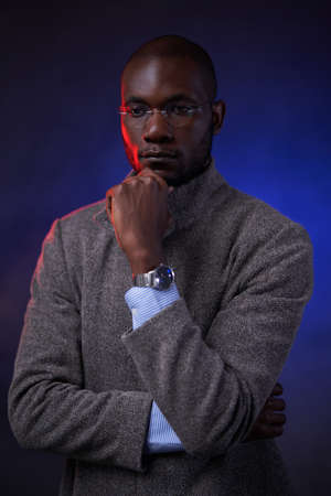 Successful African American man in gray jacket and glasses, holds hand on chin. Studio portrait on a dark background illuminated by red and blue lightの写真素材