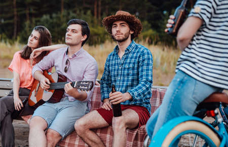 A group of friends, sitting on the pink blanket, relaxing on the beach bench, drinking beer and looking at the seaの写真素材