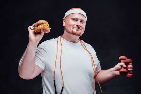 Fat men with a white headband on his bold head is holding a hamburger in one hand and two red dumbbells in other one, having an orange skipping rope on his neck, smiling at the cameraの写真素材