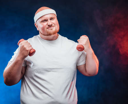 A very fat man in a white T-shirt with two dumbbells in each hand, on the contrast blue and red backgroundの写真素材