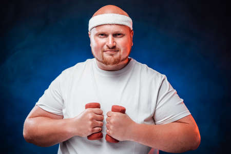 Bulky funny man in a white tracksuit holds dumbbells in his hands and looks at the camera with a smileの写真素材