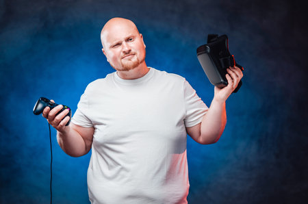 Very fat gamer in a white t-shirt holds a joystick and vr glasses in his hands, looking at the...