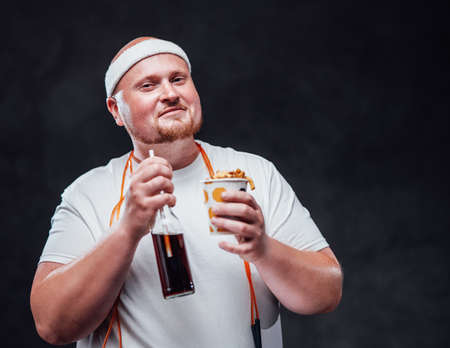 Smiling plump man in a white tracksuit, eating french fries with sauce and drinking colaの写真素材