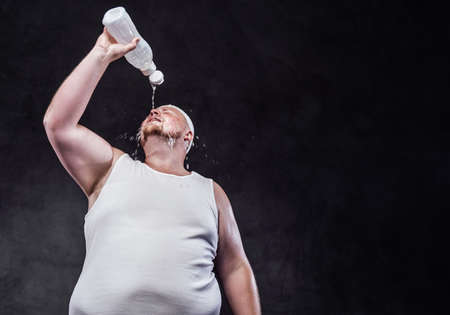 Chubby bald man, after working out, pours cold water from a bottle on his face