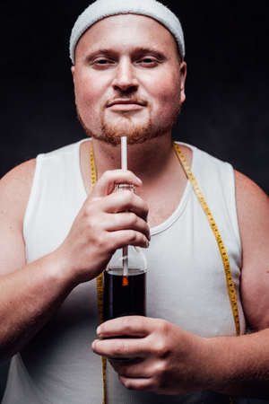 Plump man in sportswear and with a yellow measuring tape on his neck, holding a bottle in his hands,...