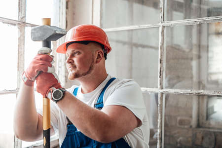 Worker, looking out the window, stands at the construction site. He is wearing a red safety helmet...