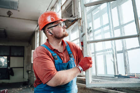 A builder in uniform and a protective helmet is carefully working with a glass window at a...
