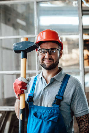 Smiling worker stands near the window at a construction site. He is wearing a red safety helmet and he holds a sledgehammer in his hands.の写真素材