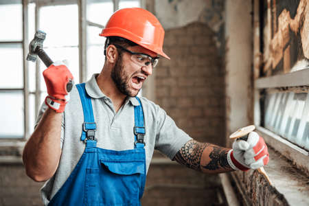 Builder destroys a wall with his hammer, screaming and looking at the detail in his handsの写真素材