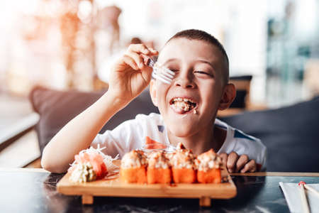 Boy enjoys eating sushi rolls in outdoor cafe, looks very funny. Bright and colorful imageの写真素材