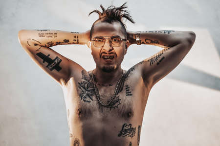 Portrait of a stylish young gangster in glasses with tattooed naked body and accessories holding...