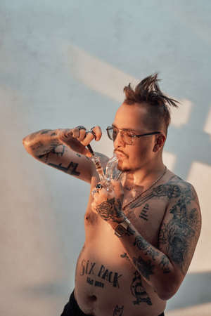 Shirtless man with tattooed body and freaky haircut lighting cannabis in the bong. Cannabis legalization conceptの写真素材