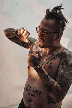 Shirtless man with tattooed body and freaky haircut lighting cannabis in the bong. Cannabis legalization conceptの写真素材