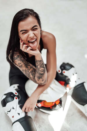 Funny smiling girl with tatoos in a black and red tracksuit, white boots and with a motorcycle helmet, posing in a white photo studio, sitting on the floor, sticking her tongue outの写真素材