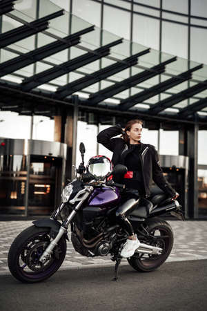 Beautiful girl in a black motorcycle jacket sits on a black sport motorbike, looking away