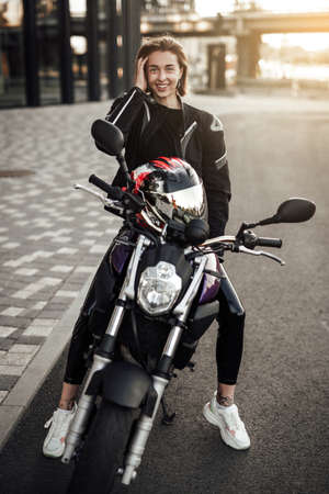 Smiling young brunette girl in a black motorcycle jacket sits on a purple sport motorbike with a red safety helmetの写真素材
