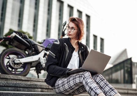 Cute and beautiful young girl is sitting next to her purple motorcycle, working at the computer. She...