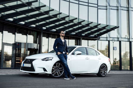 Sunny streets with skyscraper on the background. A stationary man looks away and leaning on his expensive cool car. Rich life.の写真素材