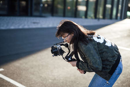 An adult woman in glasses holding a professional digital camera. The city in sunlight and photographer. Photographing hobby.の写真素材
