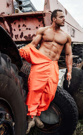 Strong man with naked torse posing with stationary old vehicle on the background. Brutal bodybuilder repairs ancient machine.の写真素材