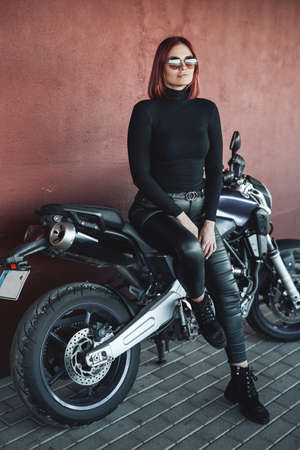 Urban motosport hobby. Beautiful woman with red hairs posing with her casual expensive motorcycle.の写真素材