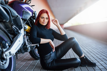 A cute young woman with redhairs sitting on the floor leaning on her bike. Custom urban motorcycle...