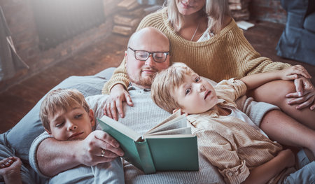 Hotels living room and joyful father and mother reading a book for their boys. Little brothers are...