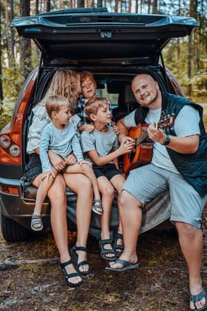 Mother and father with their boys enjoying the playing guitar and getting rest sitting in car. American summertime forest.の写真素材