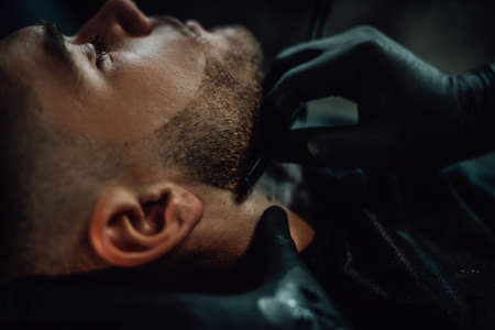 Professional stylist and hairdresser carefully shaving a beard of his client with sharp blade and black gloves.の写真素材