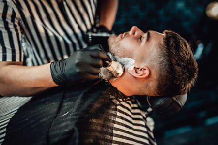 Professional hairdresser applying soap on his clients face and preparing him for cutting in retro styled saloon.の写真素材