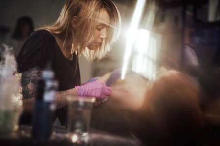 Female and caring tattoo master cleaning her clients shoulder using a doily in warm and shiny salon.の写真素材