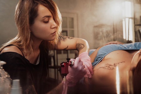 Pretty girl lying on bed and waiting while tattoo master finish drawing on her shoulder in warm and shiny saloon.の写真素材
