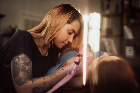 Female tattoo master holding her device and drawing waves on shoulder of her customer in illuminated warm apartment.の写真素材