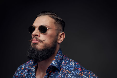 Stylish and powerful guy with huge biceps and with black beard posing in sunglasses in gray...