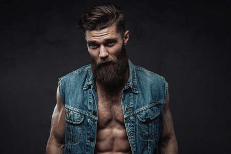 Muscular and self-confident guy with naked torso and beard with modern haircut posing in dark background.の写真素材