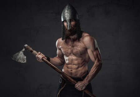 Nord barbarian with naked torso and beard in helmet posing holding his two handed axe in dark studio background.の写真素材