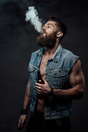 Cool and haired guy with naked torso and huge biceps posing holding bottle in arm and smoking vape in dark background.の写真素材