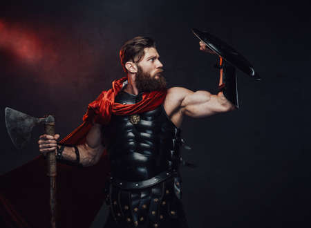 Handsome and muscular rome fighter with beard in dark armor and red cloak defends using his shield in dark background.