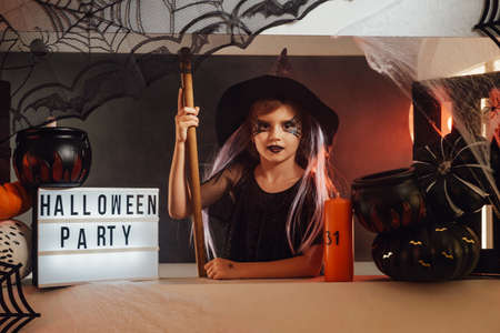 Studio room with halloween decorations and custom stand with little girl in dark clothing cosplaying witch and sitting at it.の写真素材