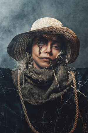 Scary woman in fashion of scarecrow with straw hat in custom clothing posing in dark smokey and atmospheric background.の写真素材