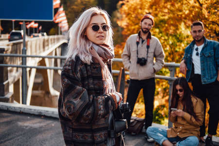 Well dressed beautiful blond posing with camera staying in foreground in background of autumn nature and her friends in daytime.の写真素材