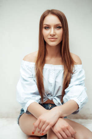Attractive and young woman with brown hairs in white shirt poses near wall on floor in white room.