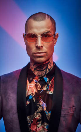 Tattooed hipster with glasses dressed in custom suit poses looking at camera with serious face in...